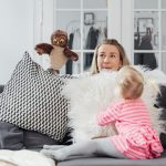 A woman and her daughter play with an owl puppet on a cozy couch in a bright living room.