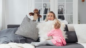 A woman and her daughter play with an owl puppet on a cozy couch in a bright living room.