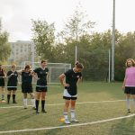 Women's soccer team practices drills on a sunny outdoor field with coach observing.